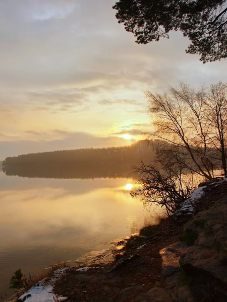 Pôr do sol de inverno sobre o lago, raios de sol laranja frios entre ramos de bétula. Derretido primeira neve no chão . — Fotografia de Stock