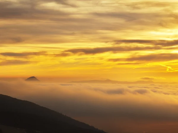 A cloud inversion is filling the bottom of valley between hills. Dark peaks of trees make contrast with yellow and orange clouds on the sky. — Stock Photo, Image