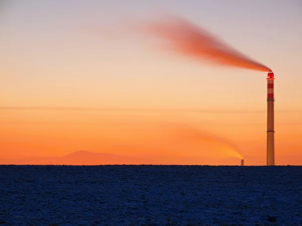 Rauchender Schornstein nach Sonnenuntergang des Wintertages, roter Himmel und dunkler Boden mit erstem Pulverschnee. — Stockfoto