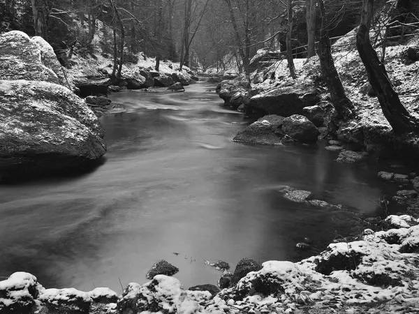 Dunkles kaltes Wasser des Gebirgsbaches im Winter zwischen großen Felsbrocken mit Schneeflocken des ersten Pulverschnees. — Stockfoto