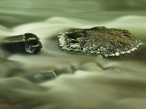Due pietre con piccoli ghiaccioli in acqua sfocata lattiginosa di torrente di montagna. Freddo clima invernale . — Foto Stock