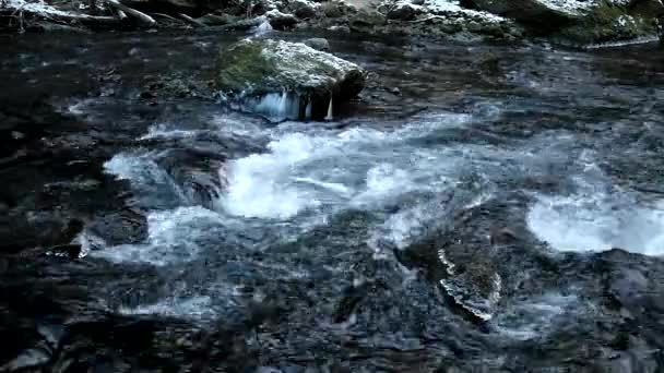 Dark cold water of mountain stream in winter time between big boulders with snowflakes of first powder snow. — Stock Video