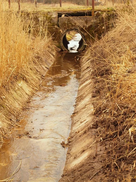 Cleared drainage channel between meadows in spring time. Water in the drain, dry old grass on banks, broken stony bridge. — Stock Photo, Image