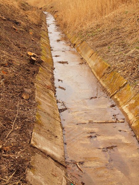 Очищенный дренажный канал между лугами в весеннее время. Вода в канализации, сухая старая трава на берегу
