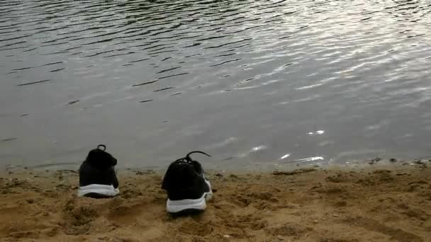 Černé a bílé tenisky na oranžové písku na břehu jezera. rozmazané vlny na vodní hladině, — Stock video