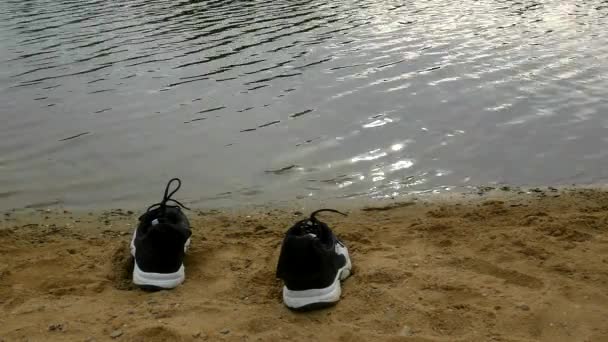 Черно-белые кроссовки на оранжевом песке озера. Размытые волны на уровне воды , — стоковое видео