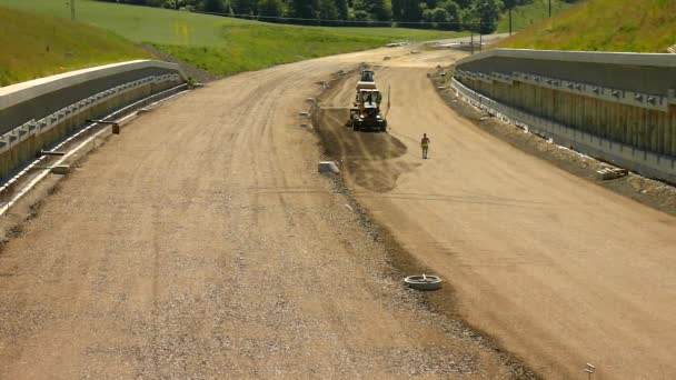 Building of new highway, scraper modify surface or gravel. Man in shorts is going. Hot sunny summer day. — Stock Video