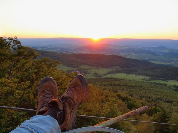 Two tired legs in blue jeans and brown leather boots with sunset at the horizon in background. Evening summer landscape. — Stock Photo, Image