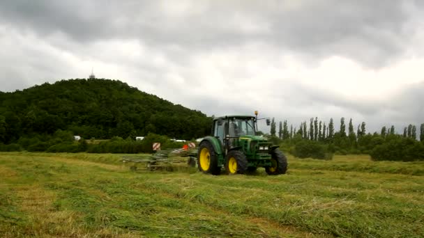 Великий зелений трактор збирає траву, вантажівка з сінозбирачем працює на лузі в сільськогосподарських угіддях. Виробництво солодощів у сільській місцевості під пагорбами. Спекотний літній день . — стокове відео