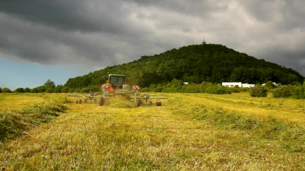 Великий зелений трактор збирає траву, вантажівка з сінозбирачем працює на лузі в сільськогосподарських угіддях. Виробництво солодощів у сільській місцевості під пагорбами. Спекотний літній день . — стокове відео