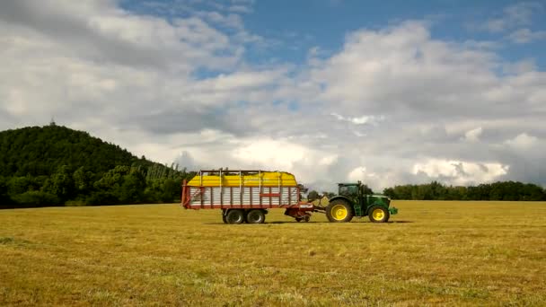 Великий зелений трактор збирає траву, вантажівка з сінозбирачем працює на лузі в сільськогосподарських угіддях. Виробництво солодощів у сільській місцевості під пагорбами. Спекотний літній день . — стокове відео