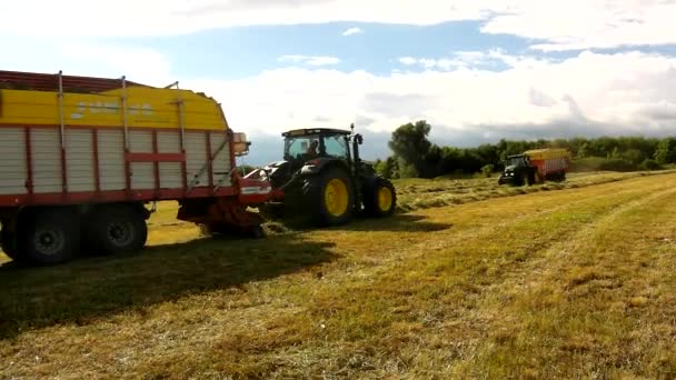 Gran tractor verde cosechando hierba, camión con heno trabajando en el prado en tierras de cultivo. Haymaking en el campo bajo las colinas. Día caliente de verano . — Vídeos de Stock