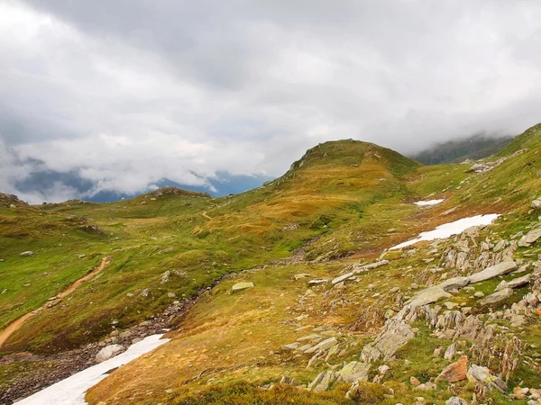 Thin footpath in snowy meadows of evening high Alpine mountains. Dark peaks in heavy misty clouds. Cold and damp end of the spring  day in Alps mountains. — Stock Photo, Image