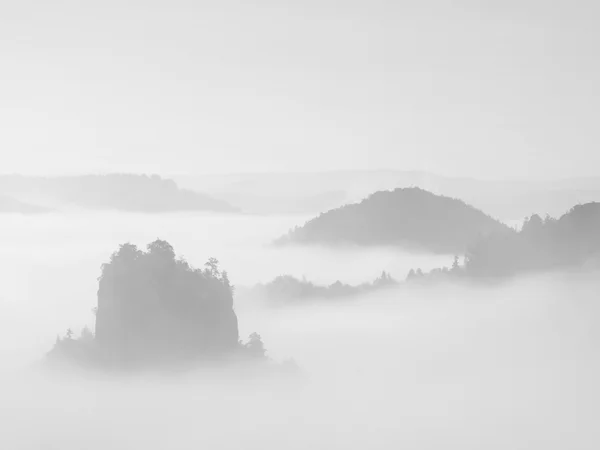Nevoeiro denso em profundo vale rochoso cheio de nuvens pesadas. picos de arenito aumentou a partir de fundo nebuloso, o sol acima. Imagem em preto e branco . — Fotografia de Stock