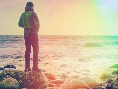 Adam deniz kıyısında taşın üstünde duruyor. Kaya üzerinde gezen gün batımını seyreder. Soyut aydınlatma, renkli işaret fişeği.