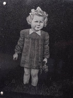 Hronov graveyard, Czechia. July 19 2022. Smiling girl with flowers in hands wears festive dress engraved as image into black granite tombstone.