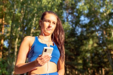 Ormanda egzersiz yapan genç ve güzel bir bayan kulaklıkla müzik dinliyor. Yaz ormanlarında kulaklıkla müzik dinleyen mutlu genç bir kadın..