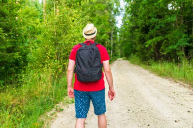 Gezgin genç adam yaz ormanlarında yolda yürüyor. Sırt çantalı genç adam sıcak yaz gününde tek başına ormanı geziyor. Turizm ve aktif yaşam tarzı kavramı..