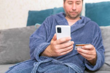 Moda ürünlerini cep telefonuyla kredi kartıyla alan genç bir adam. Online Ödeme. Bankamatik kartı tutan adamın ön portresi ve evden alışveriş için akıllı telefon kullanımı