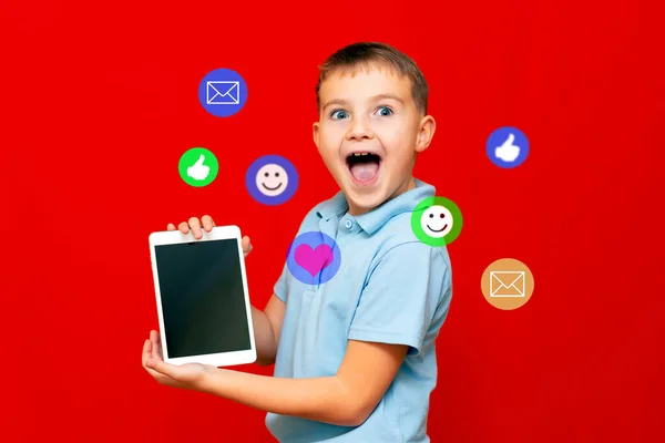 Awesome application or website. Excited 10s boy showing blank digital tablet screen and open mouth get email love heart and thumb up apps on red studio background, free space.