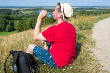 Gezgin dağlarda su içiyor. Sırt çantalı gezgin dağın tepesinde dinleniyor ve güneşli yaz gününde şişe su içiyor..