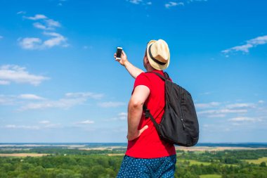 Sırt çantalı genç adam akıllı telefon kullanarak selfie çekiyor. Gökyüzü bulutlu yaz mevsimi aralığı. Manzara yaz arkaplanı. Aktif spor sırt çantalı sağlıklı yaşam tarzı konsepti..