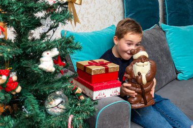 Kanepede oturan çocuk Noel Baba çikolatalı hediyeler tutuyor. Evde süslenmiş Noel ağacının yanında..