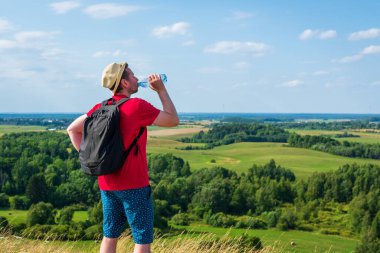 Gezgin dağlarda su içiyor. Sırt çantalı gezgin dağın tepesinde dinleniyor ve güneşli yaz gününde şişe su içiyor..