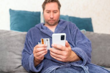 Moda ürünlerini cep telefonuyla kredi kartıyla alan genç bir adam. Online Ödeme. Bankamatik kartı tutan adamın ön portresi ve evden alışveriş için akıllı telefon kullanımı