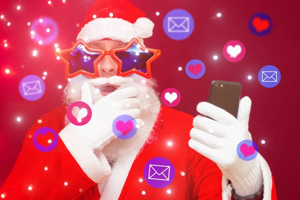兴奋的圣诞老人得到了社交媒体的关注 圣诞老人在他的手机上使用不同的社交媒体服务 红色工作室背景 — 图库照片