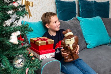 Kanepede oturan çocuk Noel Baba çikolatalı hediyeler tutuyor. Evde süslenmiş Noel ağacının yanında..