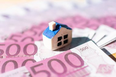 Euro banknotlarının üzerine yerleştirilmiş minyatür oyuncak evi. Gayrimenkul maliyeti, fiyatları, ev satın alma veya kiralama kavramı, hipotezi kredisi, ilgi çekici odaklanma, yakın plan. Bulanık arka plan ve ön plan.