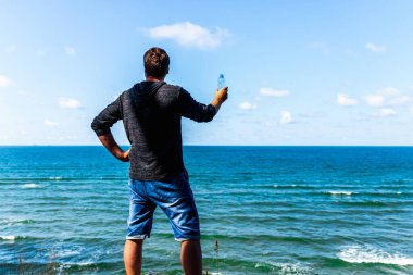 Orta yaşlı bir adam güneşli yaz günlerinde sahilde gezindikten sonra dinleniyor ve su şişesi tutuyor..