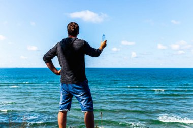 Orta yaşlı bir adam güneşli yaz günlerinde sahilde gezindikten sonra dinleniyor ve su şişesi tutuyor..