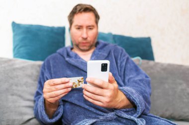 Moda ürünlerini cep telefonuyla kredi kartıyla alan genç bir adam. Online Ödeme. Bankamatik kartı tutan adamın ön portresi ve akıllı telefonu evden alışveriş için kullanmak..