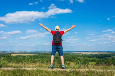 Sırt çantalı turist. Sırt çantalı, kollarını açmış gezgin. Mavi gökyüzü beyaz bulutlu yaz manzarasında duruyor.