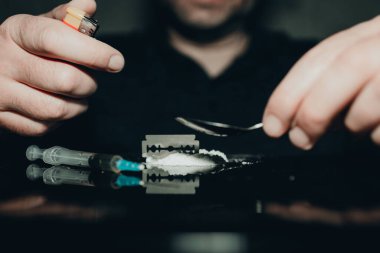 Karanlık odada uyuşturucu bağımlıları. Bağımlı, keş bir kaşık ve çakmakla uyuşturucu hazırlıyor. Beyaz toz, jilet ve şırınga. Uyuşturucu konsepti. Uluslararası uyuşturucu istismarı günü..