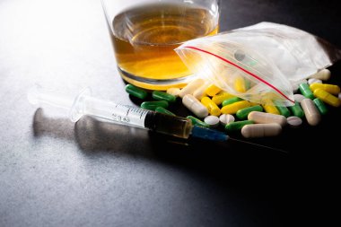 Koyu gri masada sert uyuşturucu ve alkol. Alkol hapları. Plastik kokain torbası. Masada gölgeli..