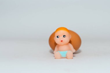 Minyatür yeni doğmuş bebek oyuncağı ve yumurta beyaz gri bir arkaplanda oturuyor. Odaklan, boşluğu kopyala.