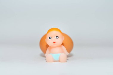 Minyatür yeni doğmuş bebek oyuncağı ve yumurta beyaz gri bir arkaplanda oturuyor. Odaklan, boşluğu kopyala.