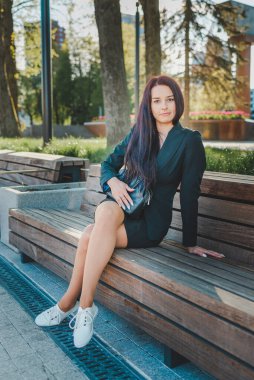 Güzel genç bir iş kadını park bankında tek başına oturuyor. Uzun saçlı esmer iş kadını, modern kıyafetiyle oturuyor, kısa eteği, ceket ve çanta eşleşmesi, kameraya bakıyor. Güzel yaz akşamı..