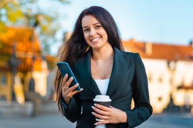 Akıllı telefonu ve kağıt bardağı olan genç bir iş kadını caddenin karşısında duruyor. Bina geçmişi bulanık. Siyah süitteki güzel bir kızın modada çekilmiş fotoğrafı. Elinde telefon ve kahve var. Yakından..