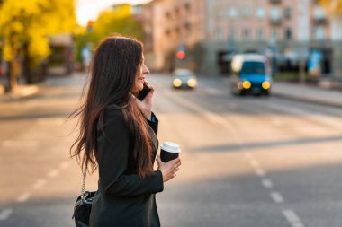 Genç bir iş kadını elinde kahve fincanıyla telefonda konuşuyor. Karşıdan karşıya geçerken geçmişi bulanık. Siyah süitteki güzel bir kızın moda fotoğrafı. Yanında telefon ve bir fincan kahve. Yan görüş..