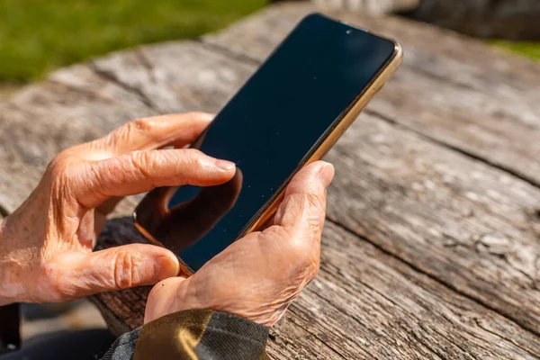 Senior woman of wrinkled finger holding and using smartphone.Old elderly woman holding phone in her hands.Closeup.In a background old wooden table,summer warm day.