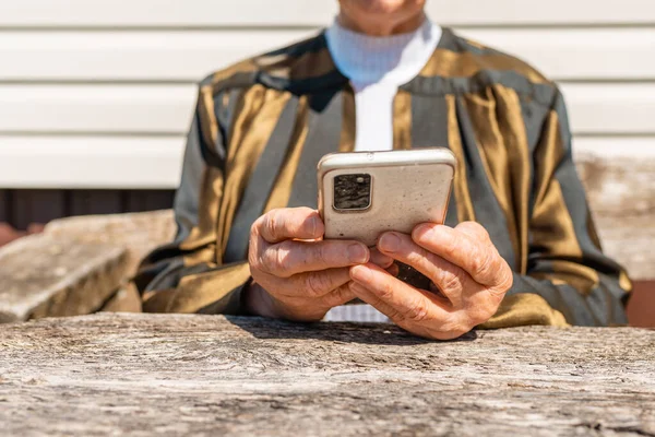 Senior woman of wrinkled finger holding and using smartphone.Old elderly woman holding phone in her hands.Closeup.In a background old wooden table,summer warm day.