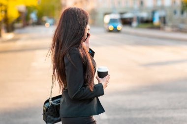 Genç bir iş kadını elinde kahve fincanıyla telefonda konuşuyor. Karşıdan karşıya geçerken geçmişi bulanık. Siyah süitteki güzel bir kızın moda fotoğrafı. Yanında telefon ve bir fincan kahve. Yan görüş..