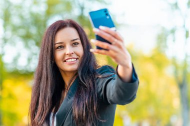 Takım elbiseli güzel bir iş kadınının portresi. İş kadını şehirde yürüyor, video görüşmeleri, selfie çekiyor, telefon ediyor. Yeni nesil iş yeri, iletişim. Bulanık yaz güneşi..