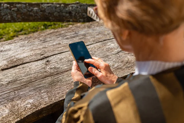 Senior woman of wrinkled finger holding and using smartphone.Old elderly woman holding phone in her hands.In a background old wooden table,summer warm day.