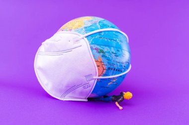 Dünya Gezegeni 'nde yüz maskesi koruyucu, minyatür oyuncak kız mor zemindeki dünya tarafından sıkıştırılıyor. Dünya tıbbi konsepti..