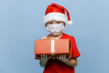 Tıbbi maske takan, Noel şapkası takan, hediye kutusunu tutan ve mavi arka planda izole edilmiş kameraya bakan mutlu küçük bir çocuğun portresi. Boşluğu kopyala